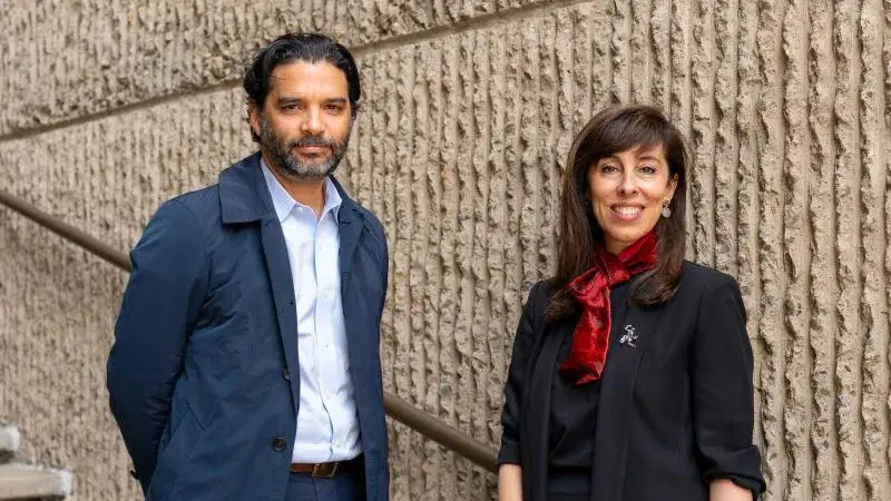 Man wearing blue jacket stands on stairs next to woman wearing black and red in front of textured concrete wall