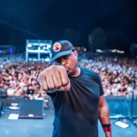 A Black man wearing a black baseball cap holds his fist towards the camera. There is a crowd in the background.