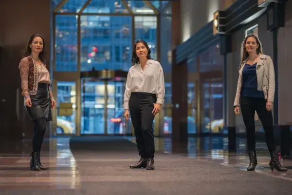 Three women stand in a space with a large window and glass door behind them