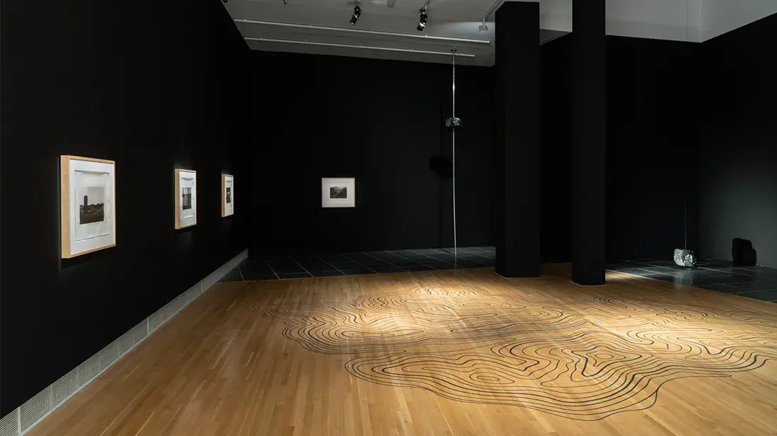 View of a gallery space with black walls. Framed black-and-white photographs hang on the wall. A topographical map is outlined on the wooden floor.