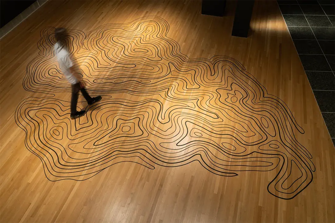 Aerial view of a person in a dimly lit gallery walking on a topographical map of a mountaintop recreated in black contour lines on a wooden floor.
