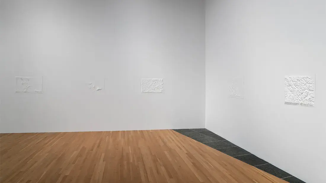 View of two white gallery walls with three rectangular white topographical maps inset into the far wall and two inset into the right-hand wall.