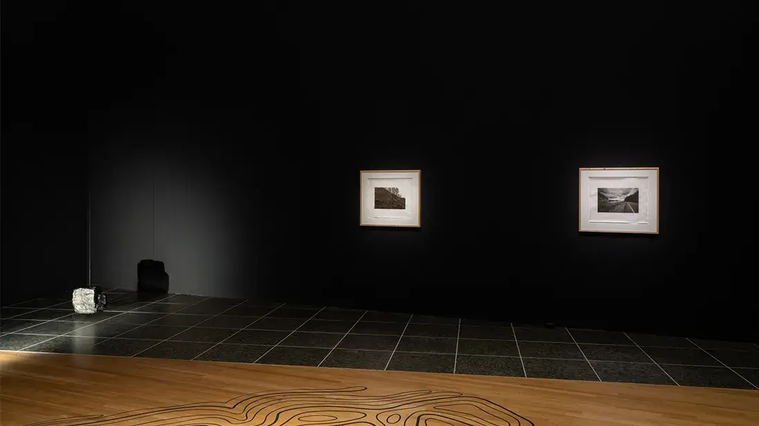 Two black-and-white photographs hang on a black wall in a darkened gallery. To the left, a square coal sculpture hangs just above the floor.
