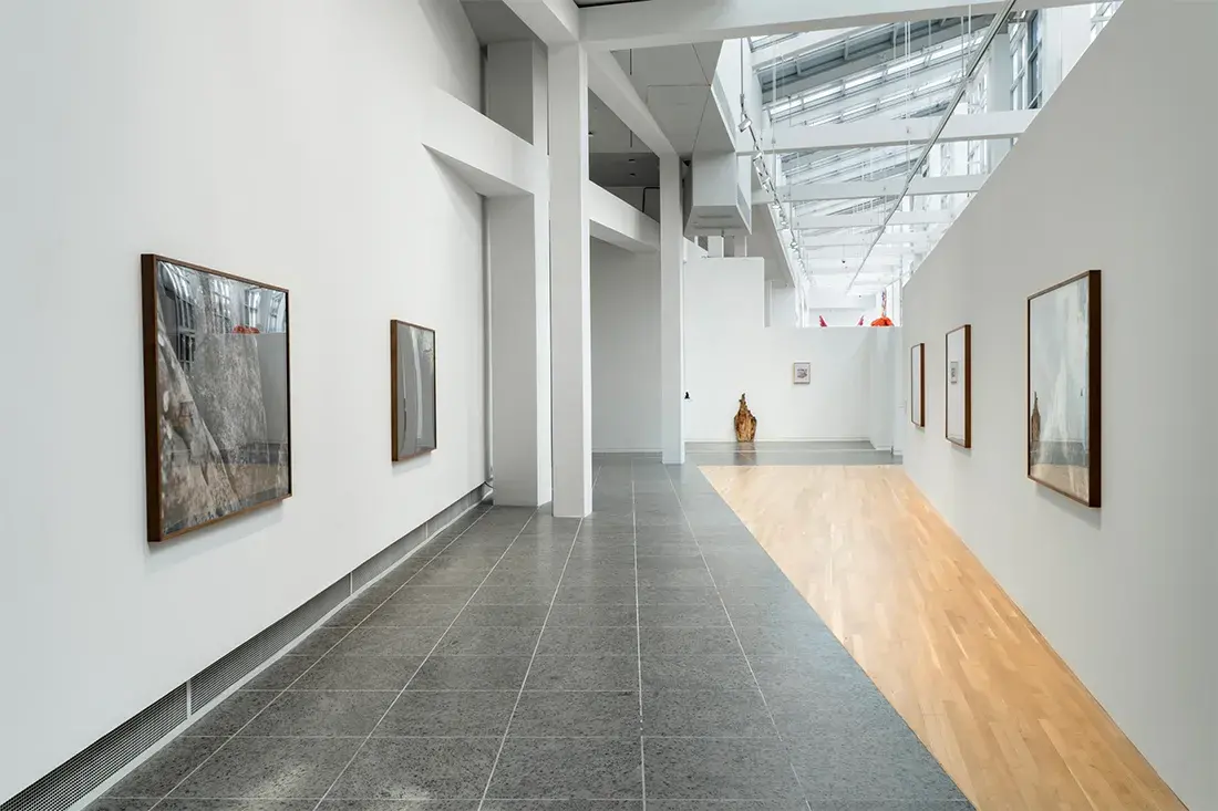 Framed closeup landscape photographs hang on opposite walls in a long gallery lit by skylights. A wooden sculpture leans on the far wall.