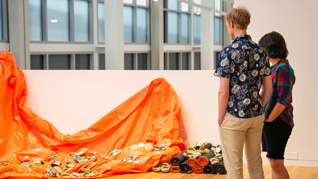 Two people look at a gallery installation. Abalone shells rest on an orange tarp draped over a low wall and on packing blankets rolled up next to it.