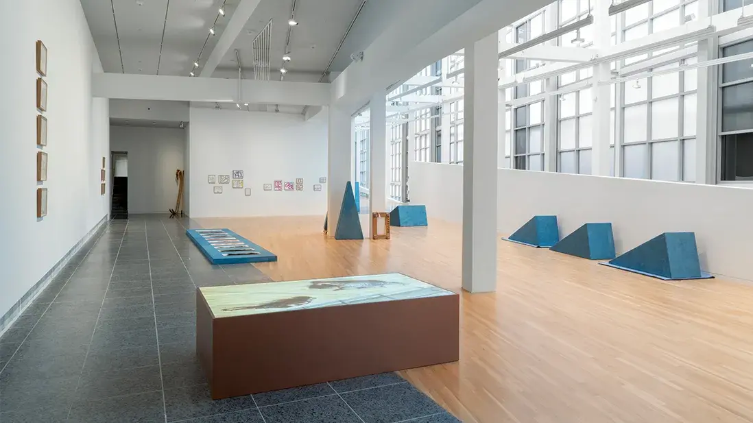 A gallery space with large windows and skylights displays blue triangular wooden sculptures, a plinth with a video projection, and framed prints.