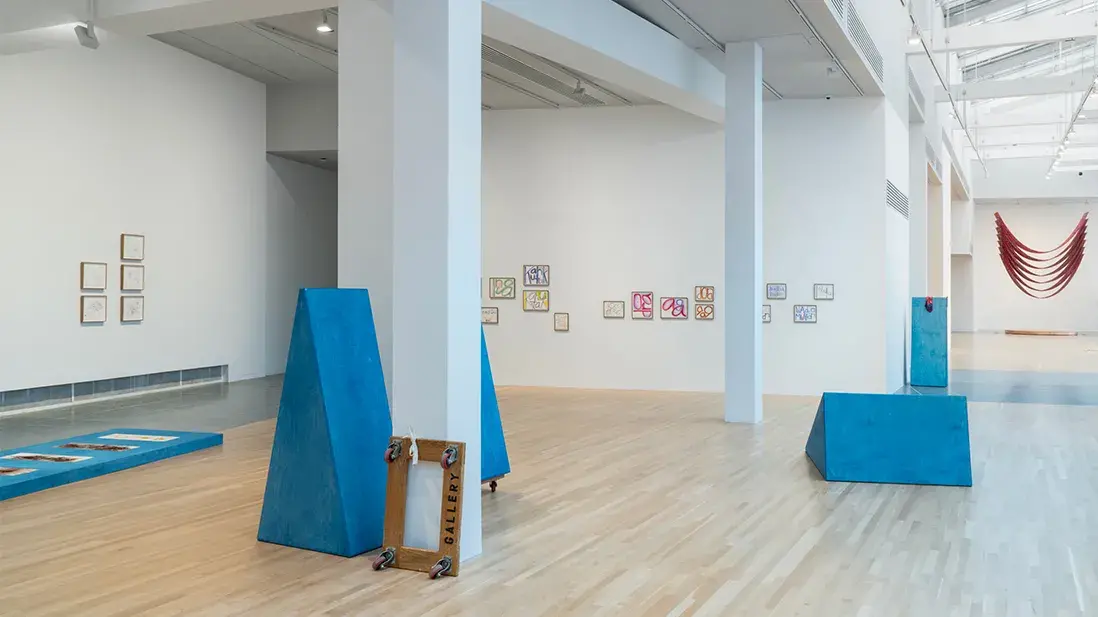 A gallery space with large windows and skylights displays blue triangular wooden sculptures, a furniture dolly, and framed prints.