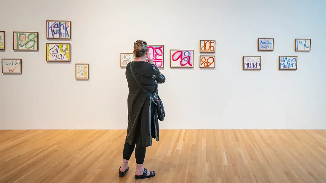 A person looks toward a gallery wall with fifteen framed paintings of abstracted, brightly colored, script-style letters and words. 