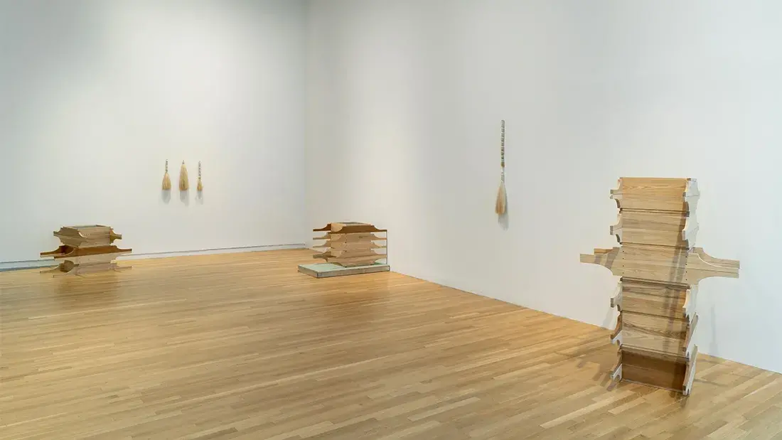 Three stacks of wooden, handled sifting trays sit on a wooden gallery floor. Three horsehair bundles hang on one wall, and one on the adjacent wall.