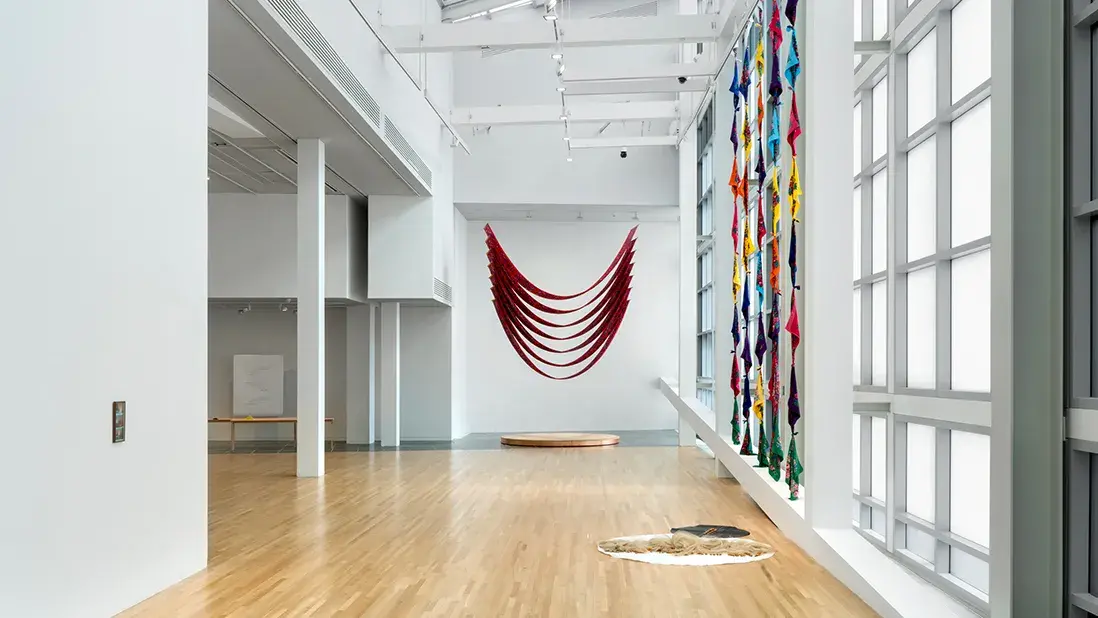 Rows of red scarves hang in a U-shaped above a circular platform. Knotted colorful scarves hang in front of windows near a tarp displaying horsehair.