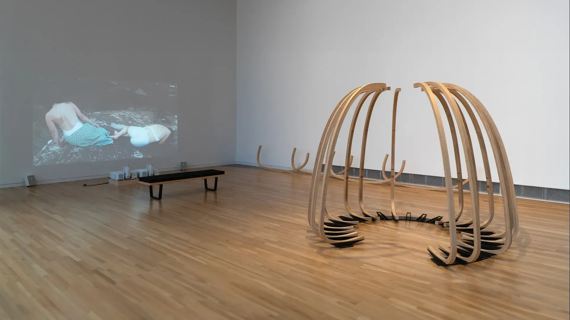 A tall, semicircular sculpture of fifteen curved wooden ribs on a metal base sits near a video projection depicting two figures reclining on a rock.