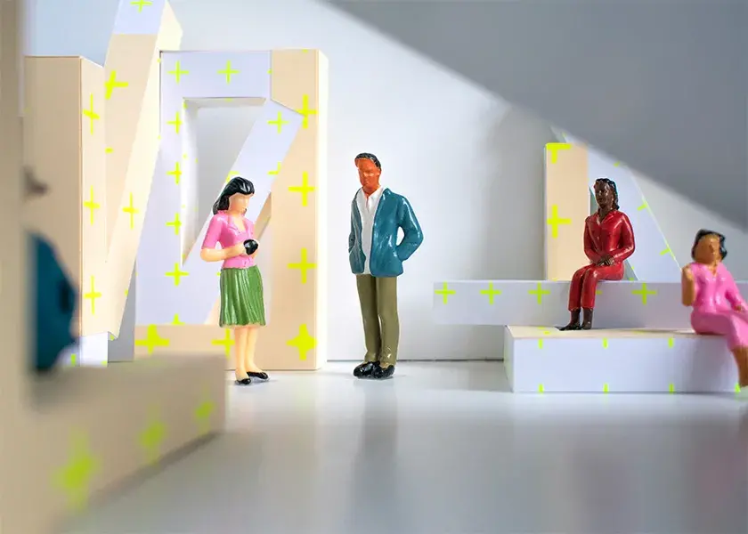 Figurines sit and stand in a white space featuring large, white and beige geometric, architectural elements with neon yellow plus signs on them.