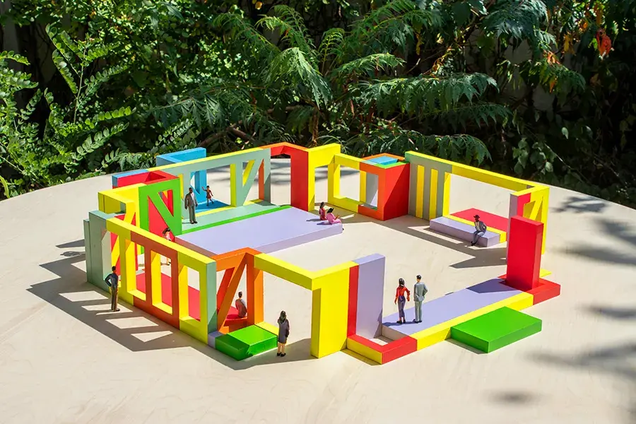 An architectural mock-up. Colorful, geometric design elements are arranged in a square within a courtyard. Miniature figures stand on the blocks.