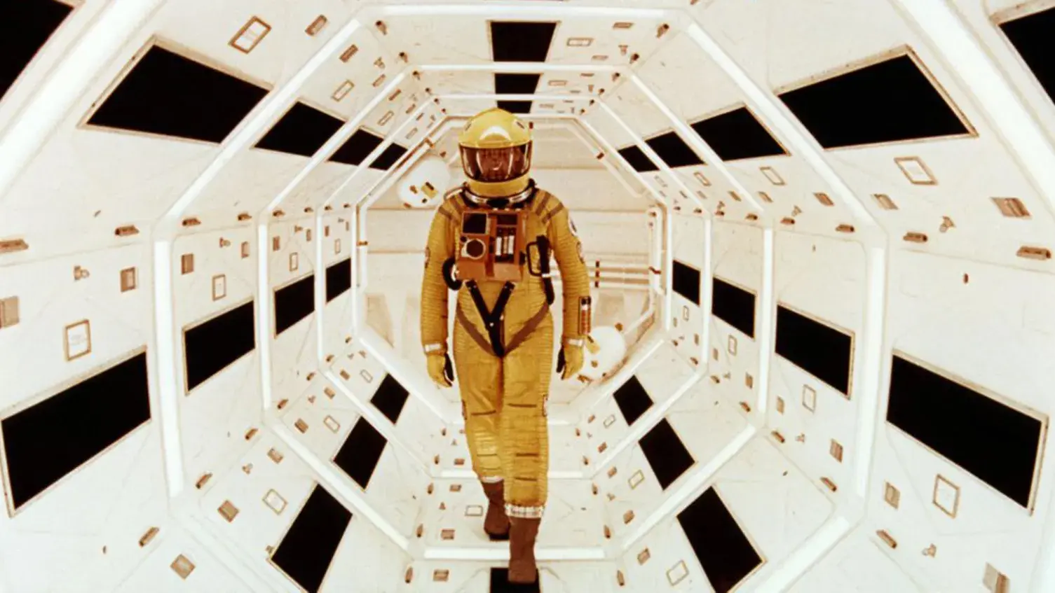 An astronaut in yellow walks down a brightly lit futuristic hallway.