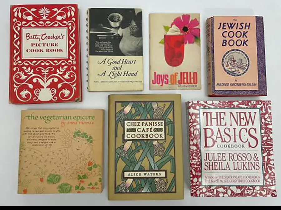 A group of 7 cookbooks lay flat. They each have different designs and contain recipes from different cuisines.