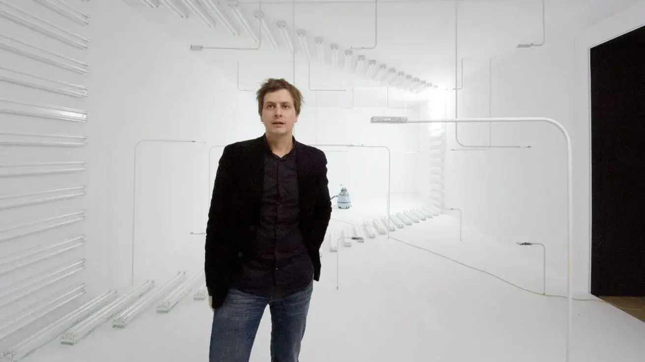 A white man wearing a black jacket and jeans stands in a white room with unlit white lights behind him in a pattern