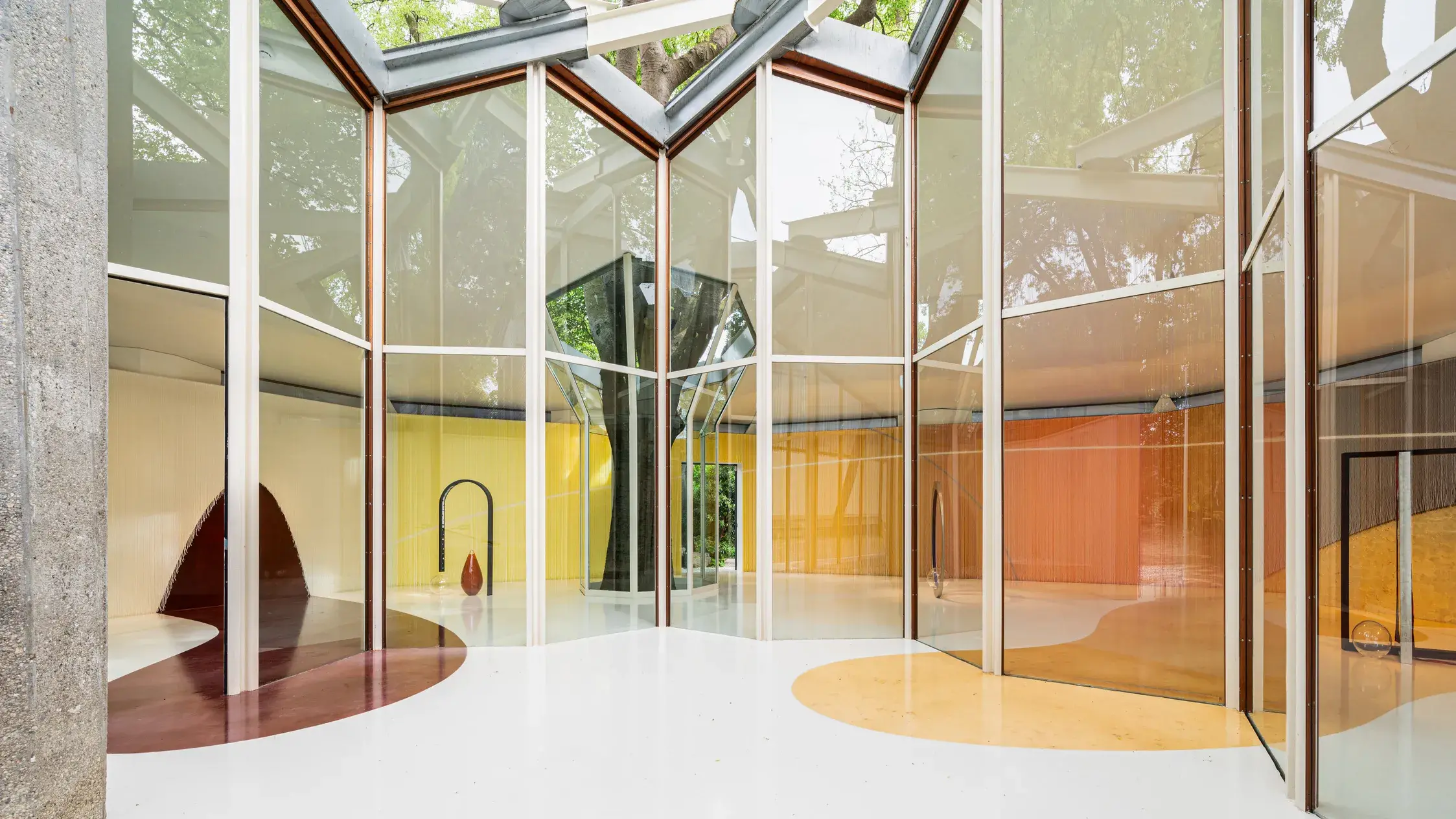 A look into an architecture building with tall angular glass windows. A large brown abstract shape is painted on the left side of the floor and bleeds up into the wall. A large orange-ish yellow abstract shape is painted on the right side floor and wall area.