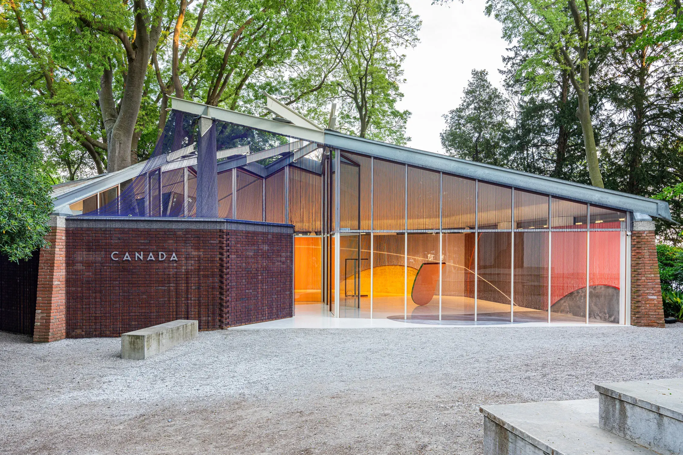 An architectural building with a slanted roof, brick detailing on the lower left side and large vertical windows showing a gallery exhibit inside.