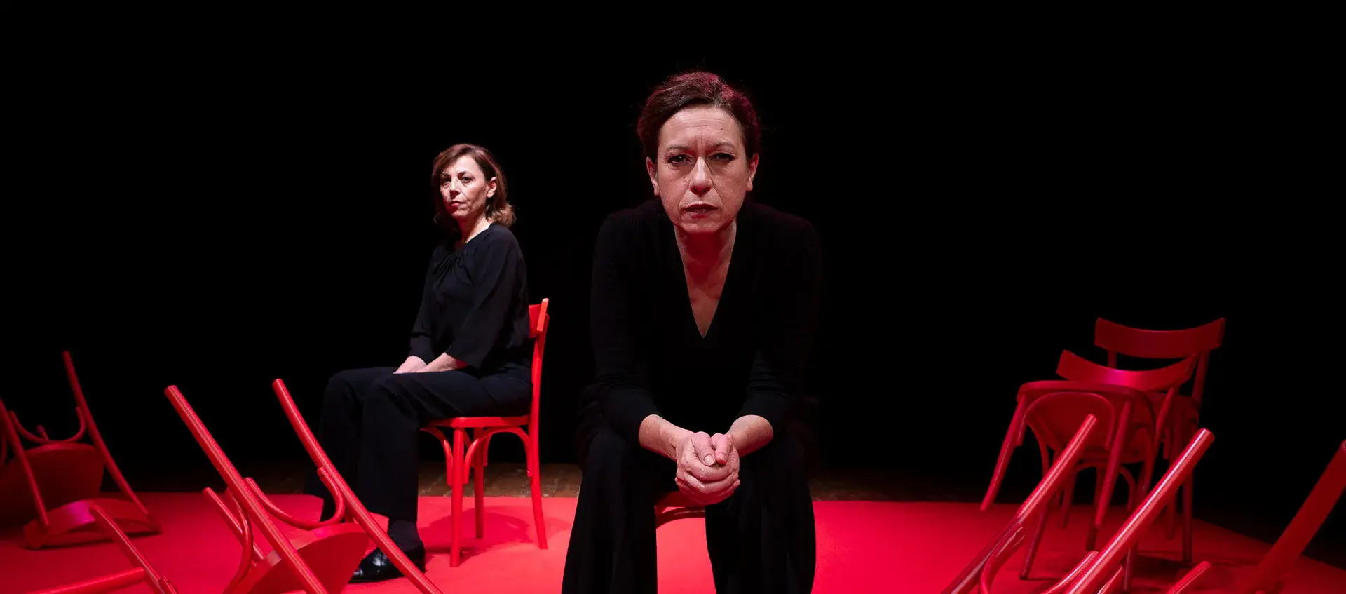 Two women sitting in red chairs in a dark room stare intently at the camera.