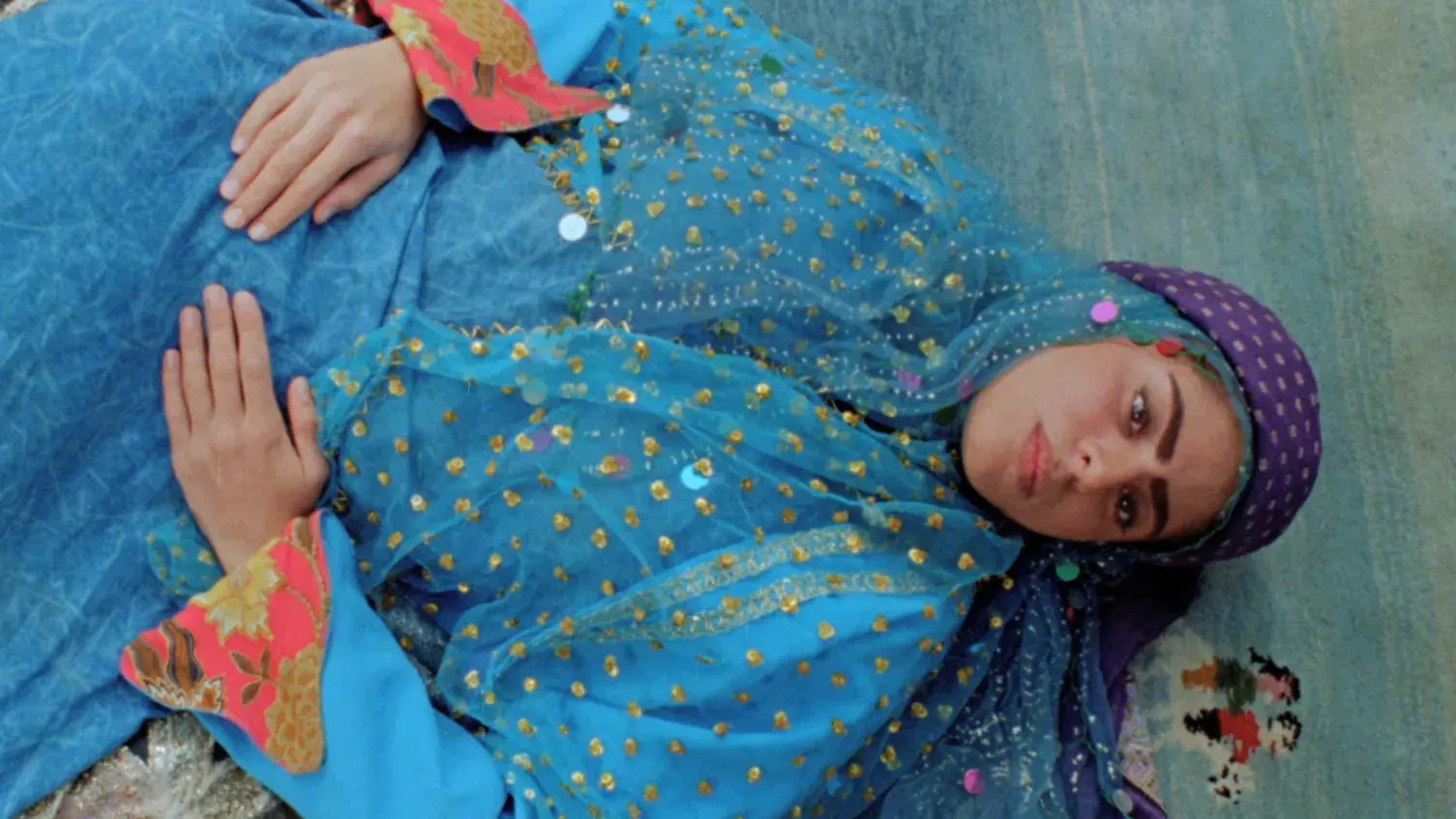 A woman in blue Iranian garments lying on the ground looking toward the camera.