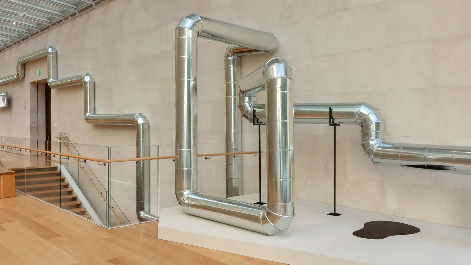 An industrial steel pipeline hangs on a wall above a museum stairwell. It frames architectural elements and ends on a plinth with a pool of oil on it.