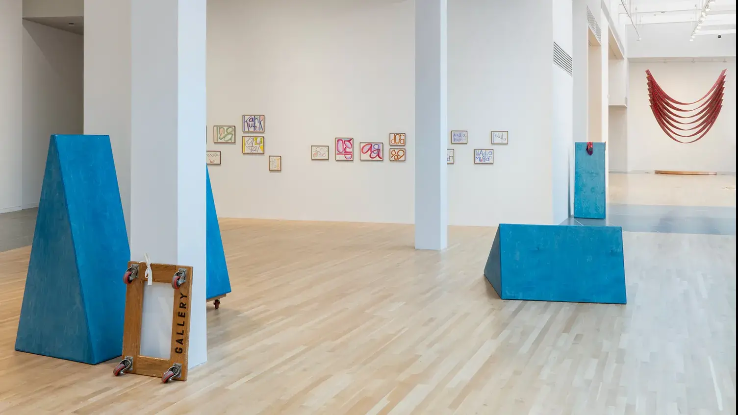 Gallery view of four freestanding triangular blue sculptures, prints displayed on a riser and the walls, and a hanging, U-shaped, red fabric work.