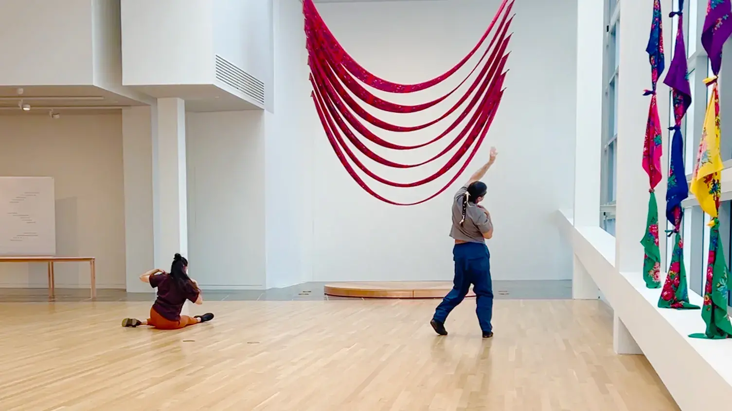 Two performing artists dancing in a gallery. One artist is seated on the ground elbows bent, hands on shoulders. The other artist stands on the right side of the image stretching their left arm towards the right window.