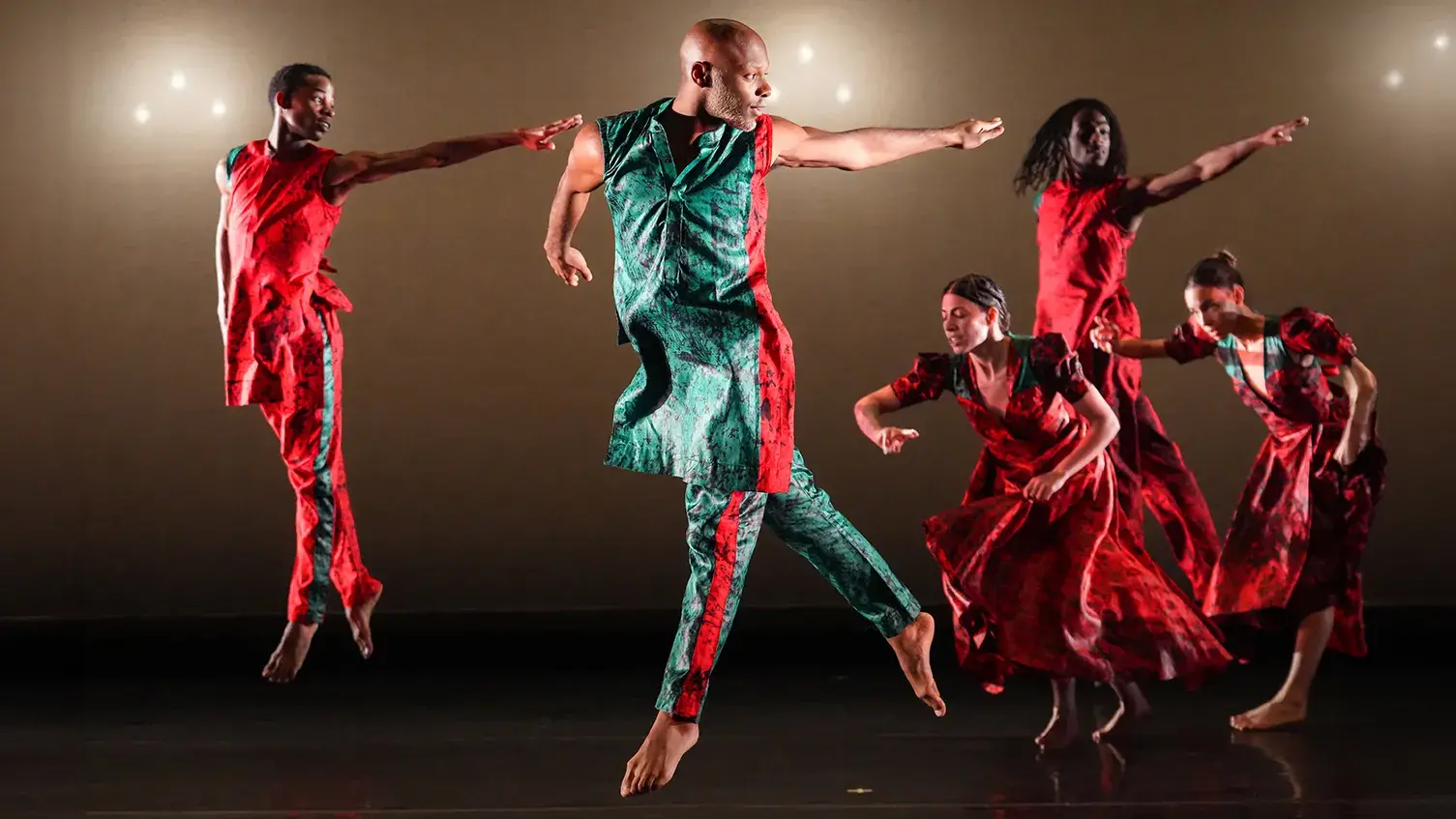 Five dancers, wearing red and green attire, perform and jump on stage.