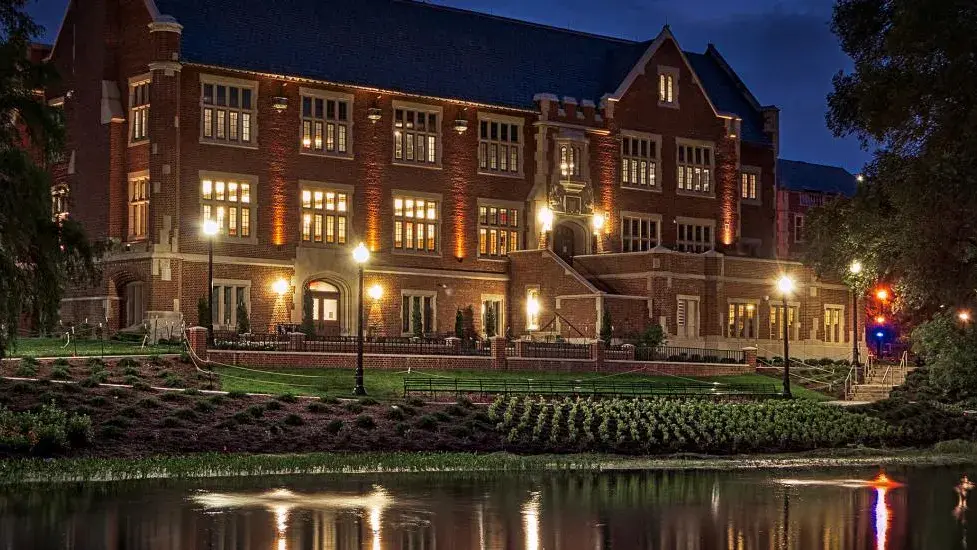 Large brick building with many lights on inside in front of landscaping and a pond