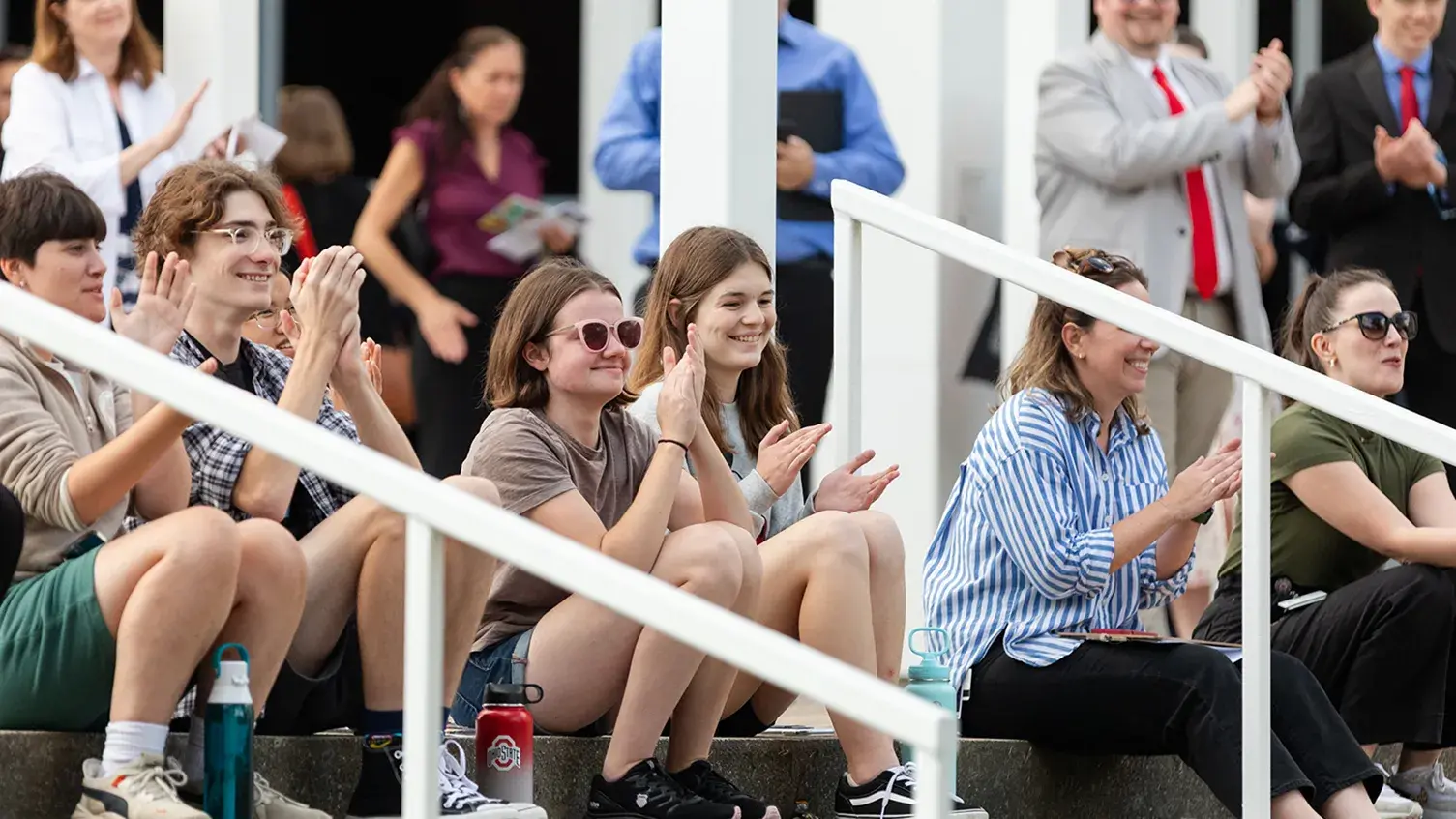 Students and community members sit on stairs clapping and smiling.