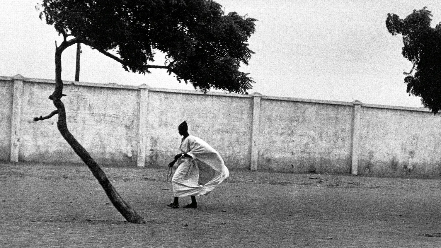 Figure with a billowing garment walking along a wall near trees.