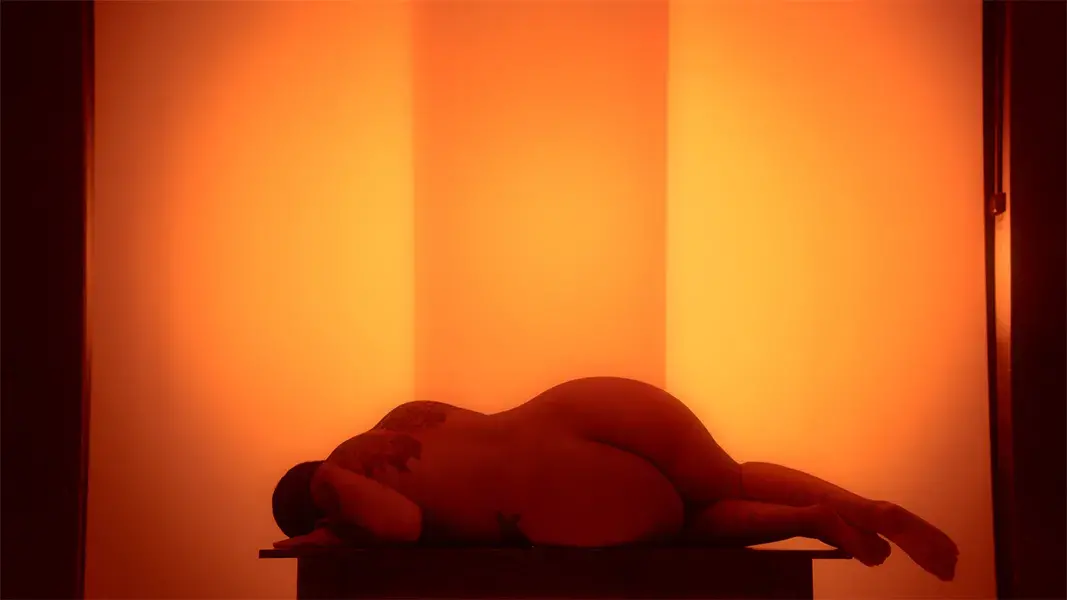 The artist lies naked on a table against a backlit amber background. She is on her side, facing away from the camera, with her head turning downward. 