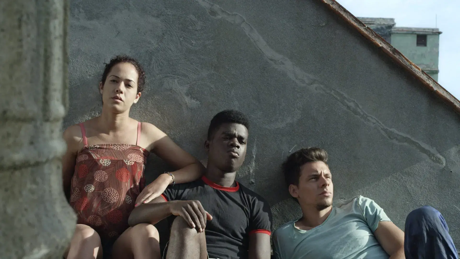 three young people sit against a gray wall outdoors