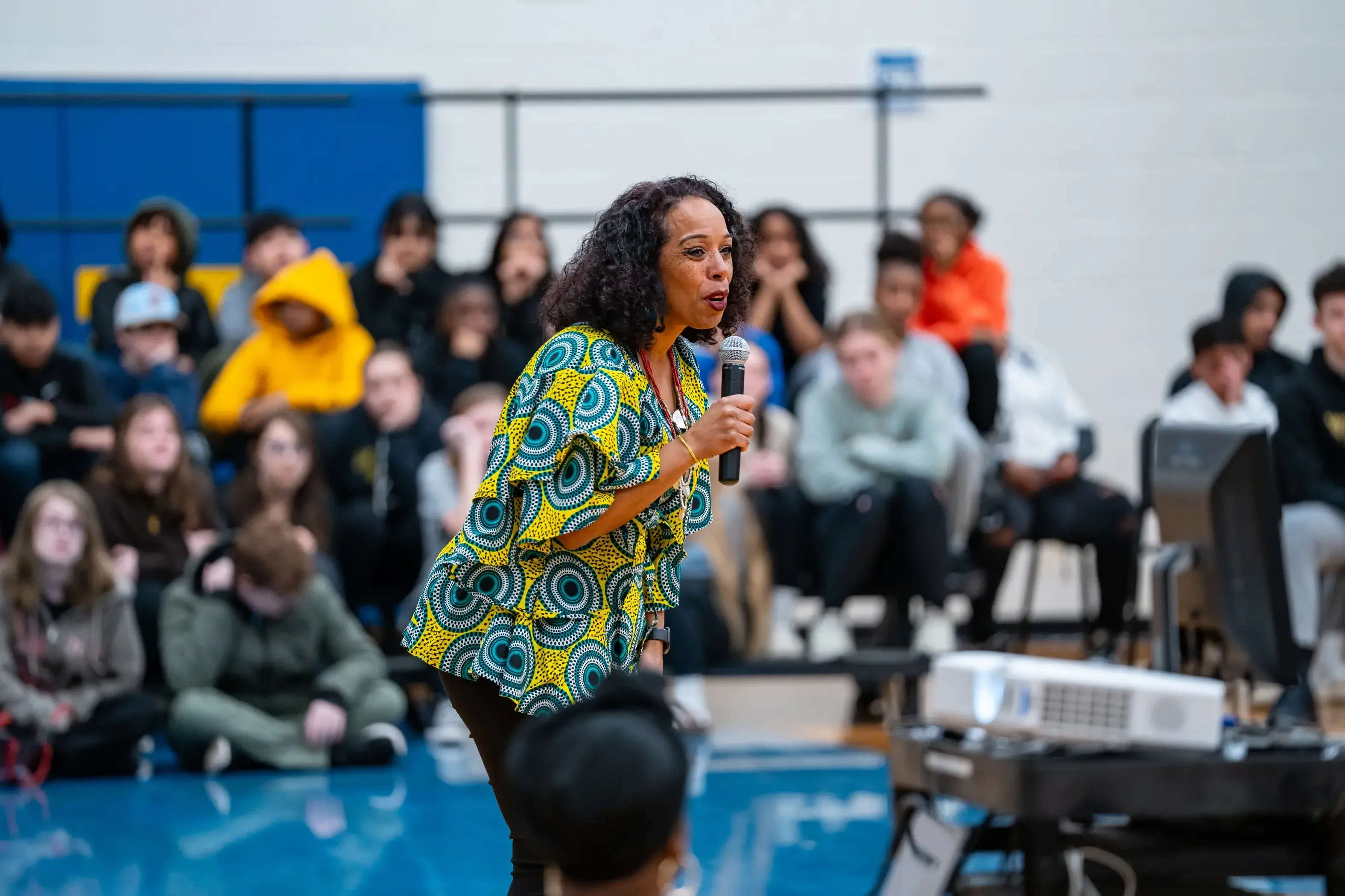 Nyama McCarthy Brown, a Black woman with dark hair, speaks into a microphone in front of a group of young people sitting on risers