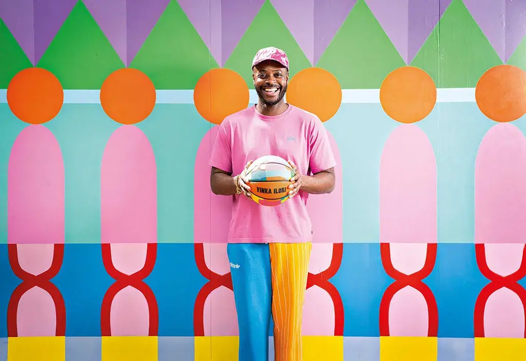 A smiling dark-skinned man holding a multicolored basketball stands in front of a multicolored patterned wall