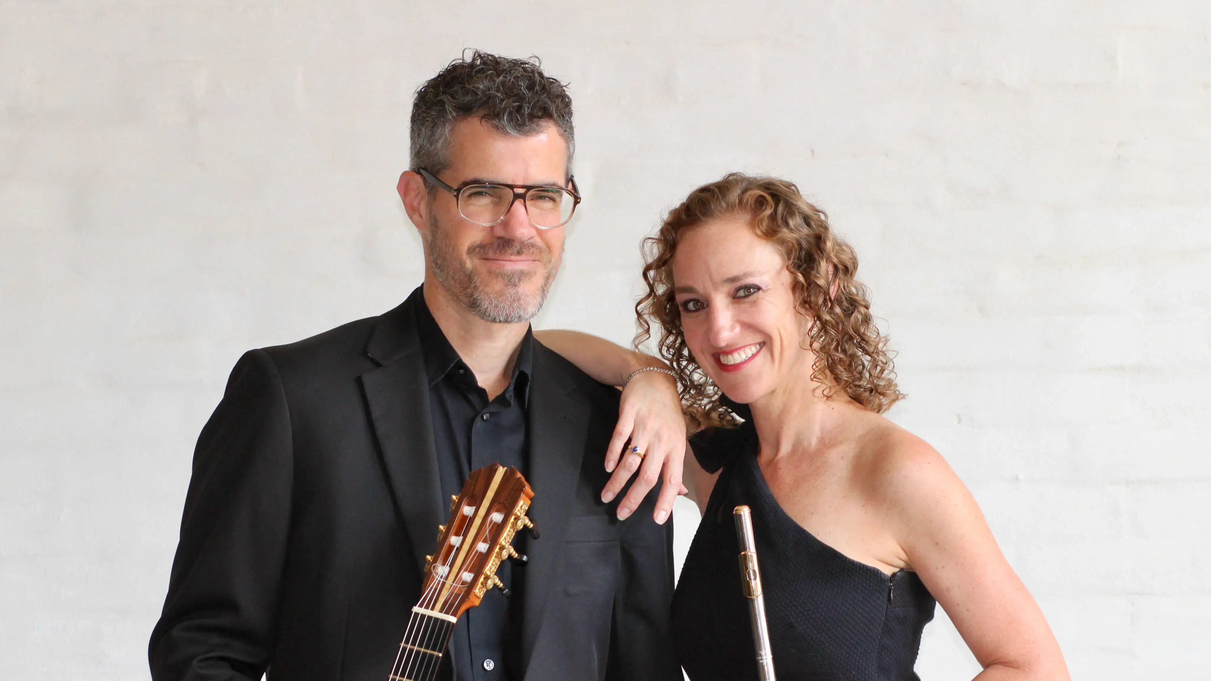 A woman with brown curly hair holds a flute and leans on a man with dark gray hair wearing glasses and holding a guitar.
