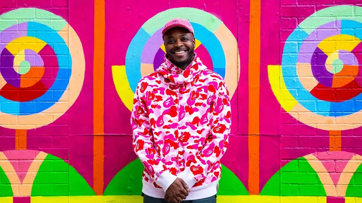 A smiling dark-skinned man wearing a hat stands in front of a brightly colored painted wall