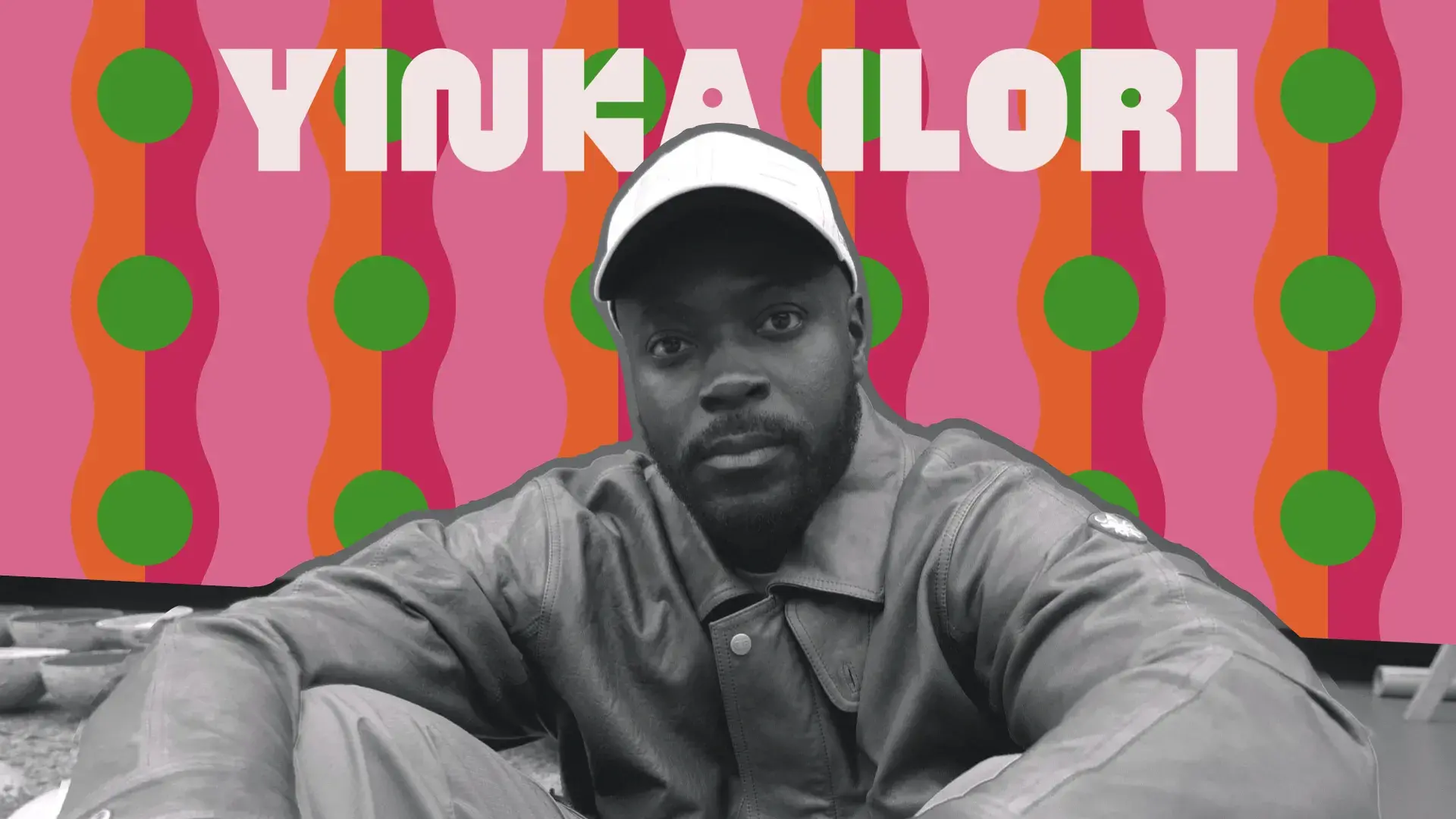 A black and white photo of a man wearing a hat sitting in front of a patterned colored background of pinks, orange and green. His name, Yinka Ilori, appears at the top of the image.