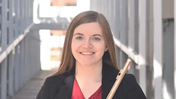 A young woman with straight brown hair holds a flute in front of her and smiles at the camera