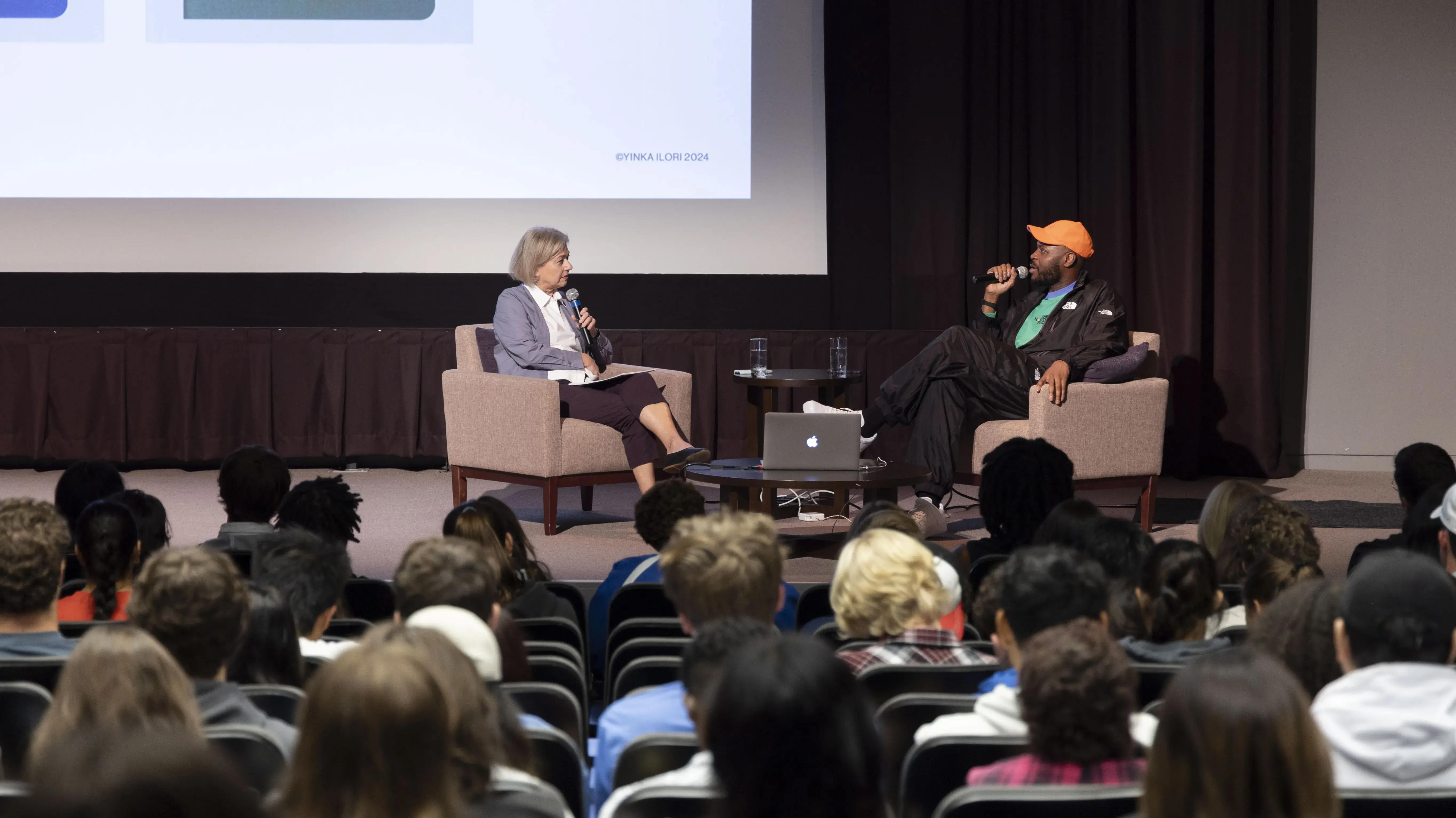 A Black man wearing an orange ball cap sits on a stage in front of theater seats full of people speaking to a white woman with blonde hair. 