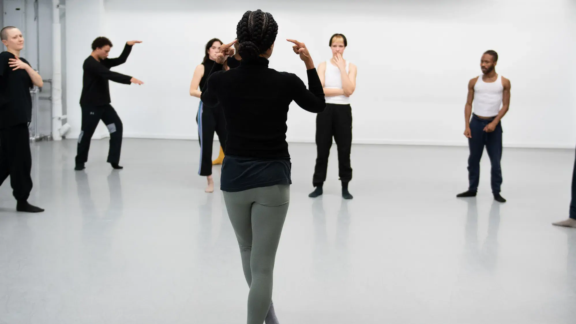 A Black woman wearing a black shirt and light green leggings stands with her back to the camera, gesturing to a small group of people facing her in a circle