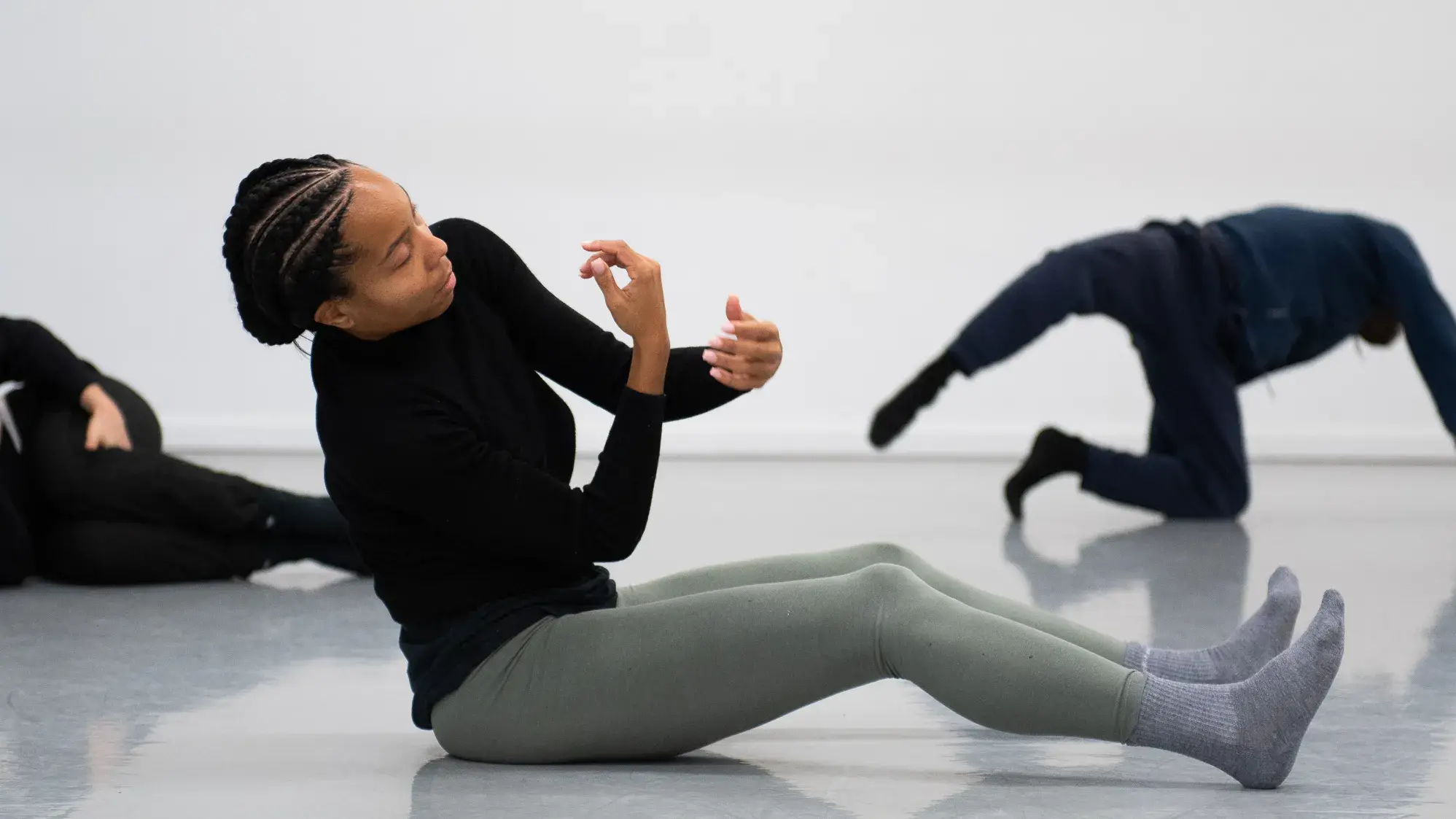 A Black woman wearing a black shirt and light green leggings sits on the floor and is in movement. Two other dancers wearing black are in the background.