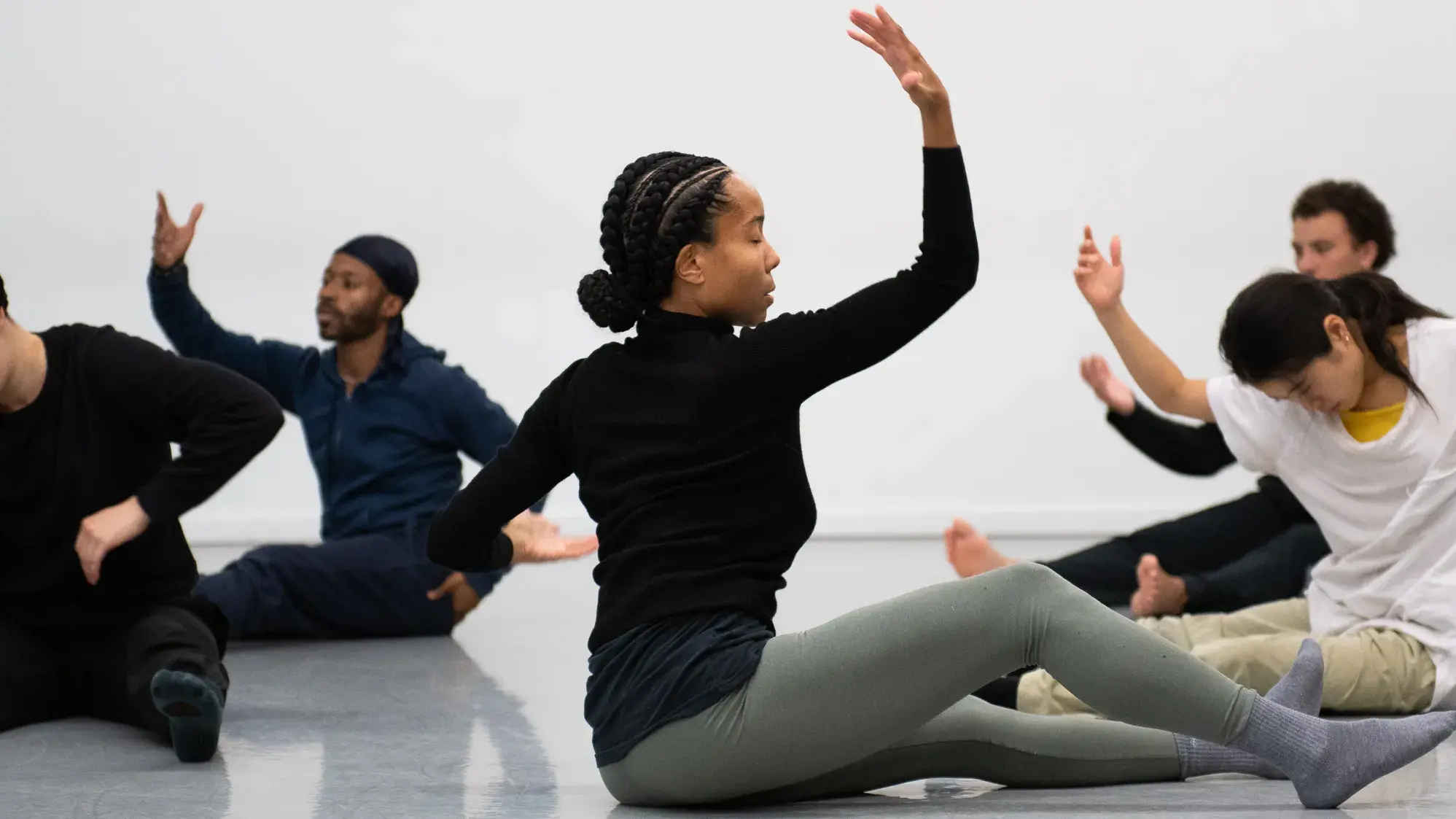 A Black woman wearing a black shirt and light green leggings sits on the floor and is in movement. Several other dancers are in movement near her.