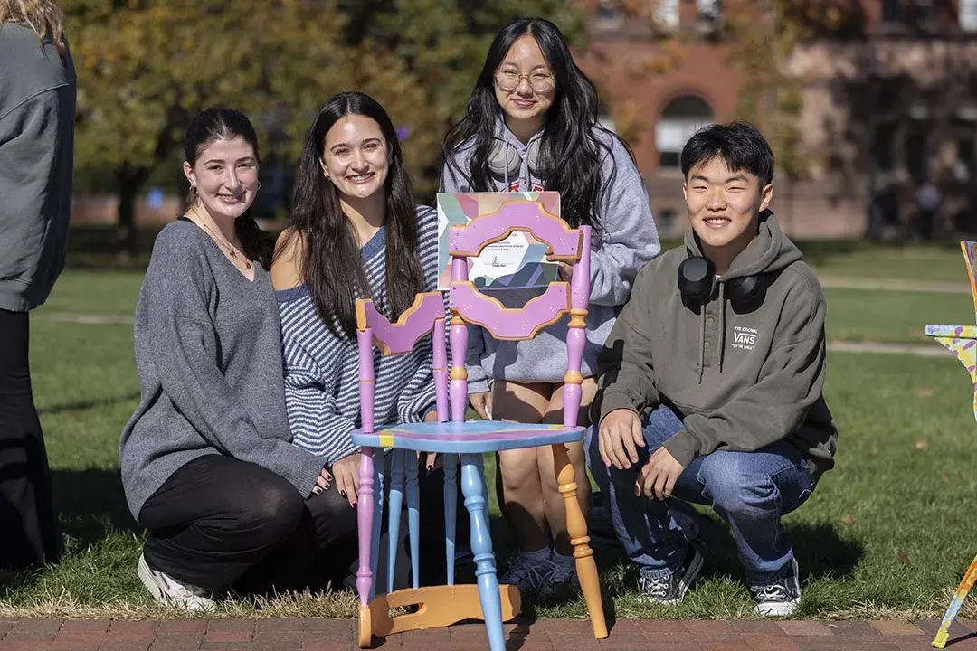 Four young people kneel behind a multicolored chair. The chair has an arm on only one side and there is a piece on the bottom of the chair that typically would serve as the back.