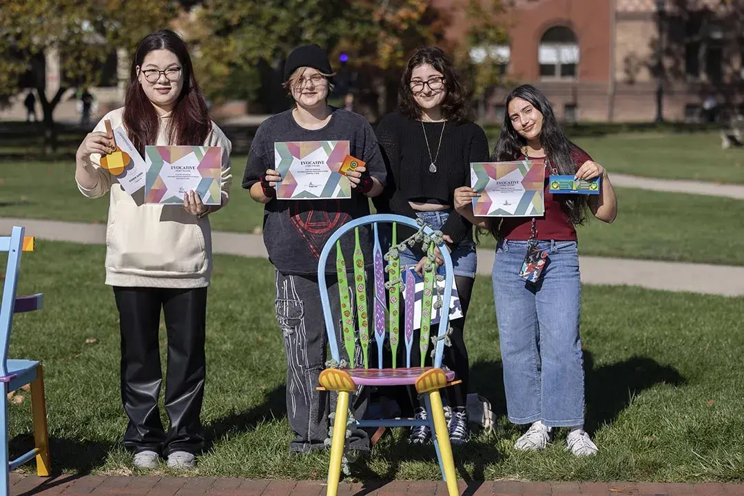 Four young people stand behind a multicolored chair with patterns on the back pieces, some of which have been cut away.