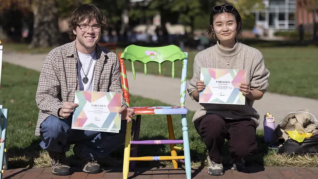 Two young people kneel next to a multicolored chair. Pieces of the back have been cut away.