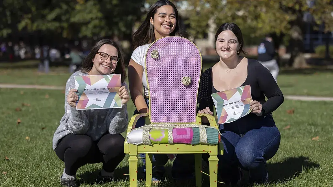 Three young people kneel behind a multicolored chair with lime green legs, a purple back and a fabric-covered seat of pinks, greens, and white