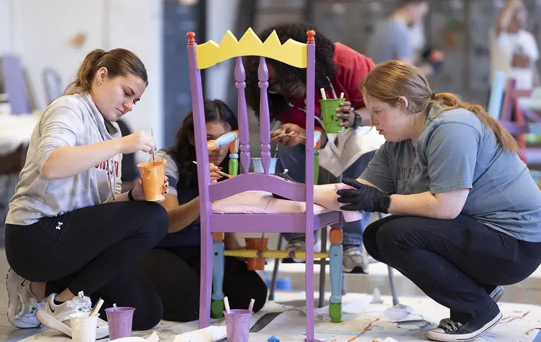 Several young people gather around a chair and are painting it in several bright colors, like purple, pink, and yellow.