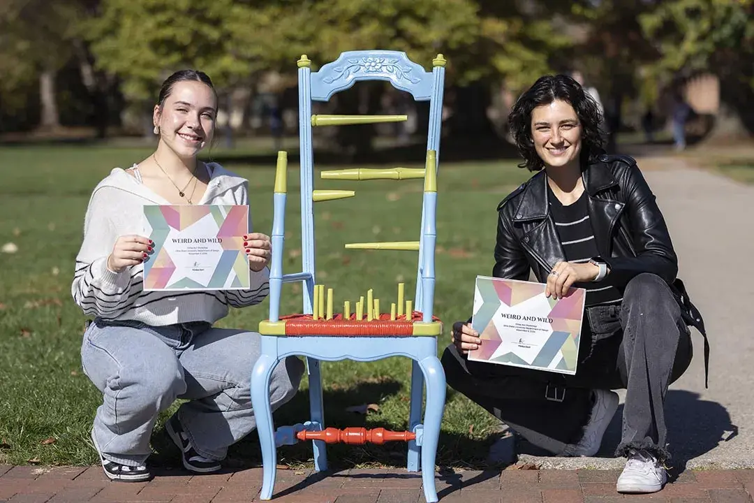 Two young people kneel next to a multicolored chair. The chair has no arms, the pieces of the chair back do not go all the way across, and there are vertical pieces of varying heights sticking up from the seat.