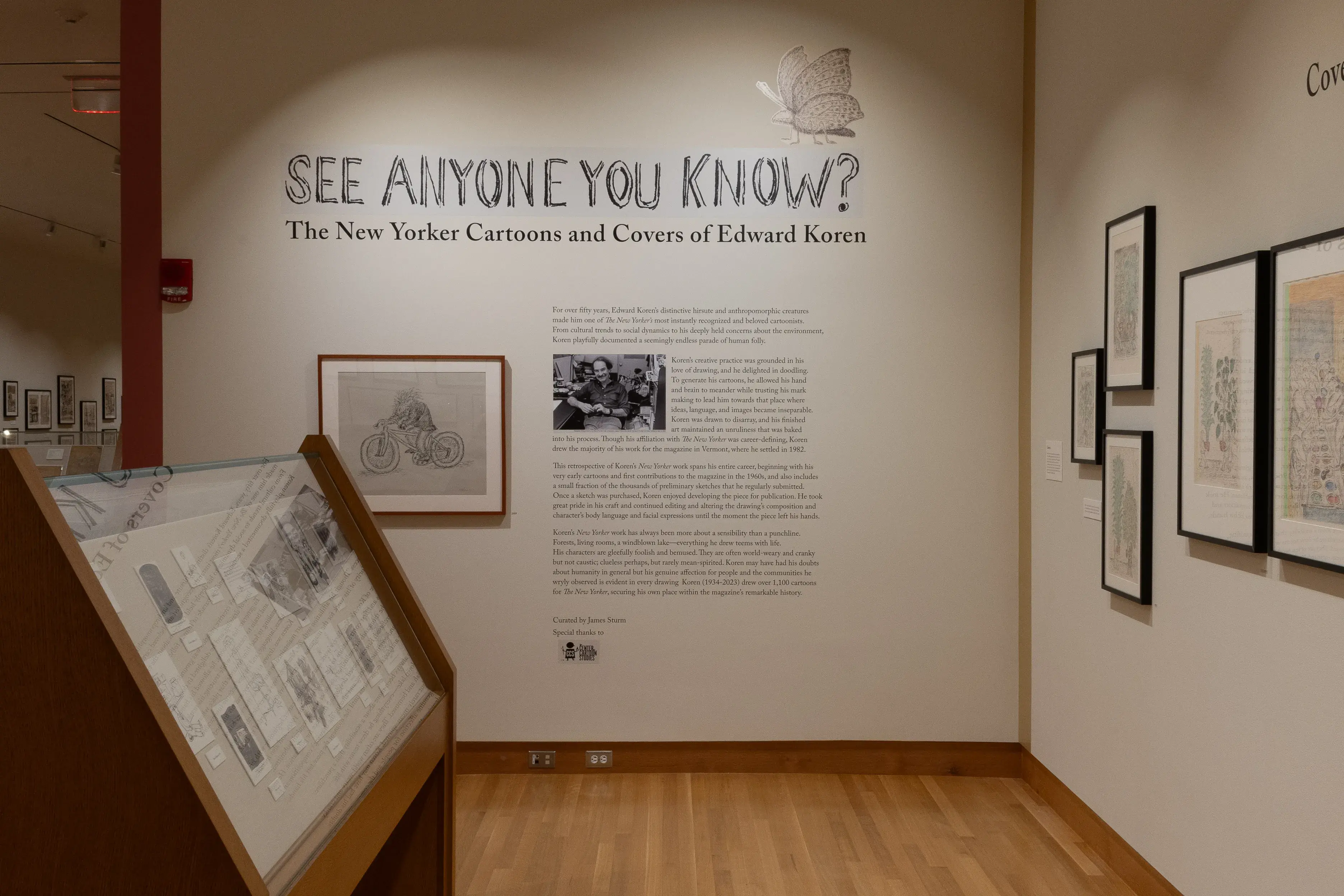 Installation image of a gallery exhibition. The title of the exhibition is on the wall, "See Anyone You Know?" with text below it. There are framed drawings on the wall and more drawings in a glass case in front.